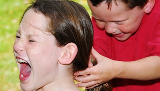 A boy in a red shirt laughing and pulling a brown-haired and freckled girl's ponytail 