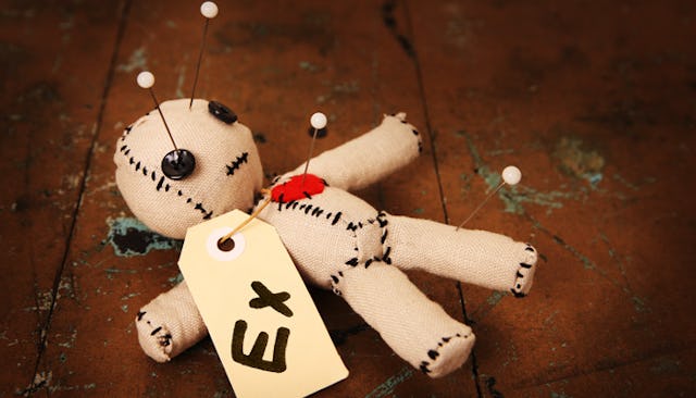 A white doll with black stitches, black buttons for eyes and a tag with text "EX" stabbed in a red h...