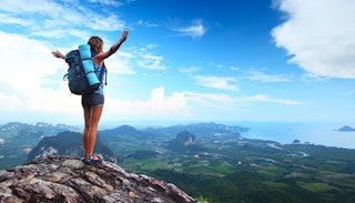 A woman in her 20s in shorts and a blue tank top with a backpack standing on a top of a hill with op...