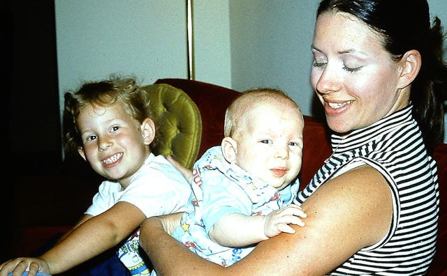 A 1970s mom in a striped sleeveless turtleneck holding a toddler and baby in her living room