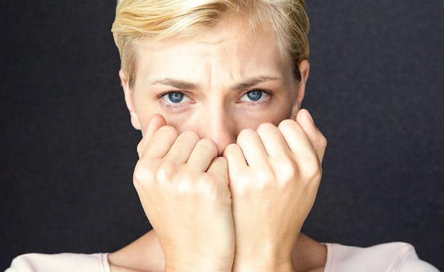 A sad looking short-haired blonde woman with blue eyes, with her hands covering her acne scars
