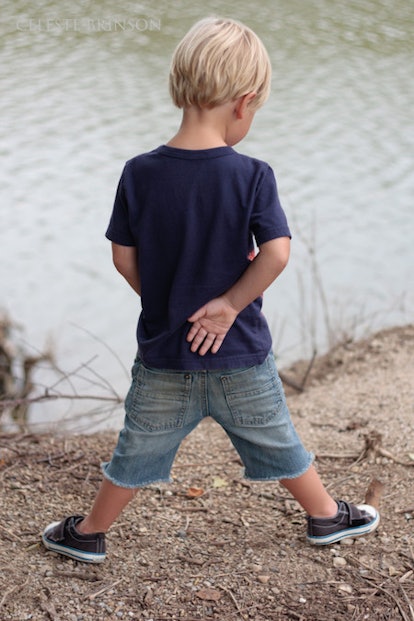 A blonde boy in a navy shirt and blue denim shorts standing and looking into a lake