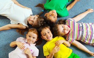 Five colorfully dressed children laughing as they lie in a circle on the floor with their heads toge...