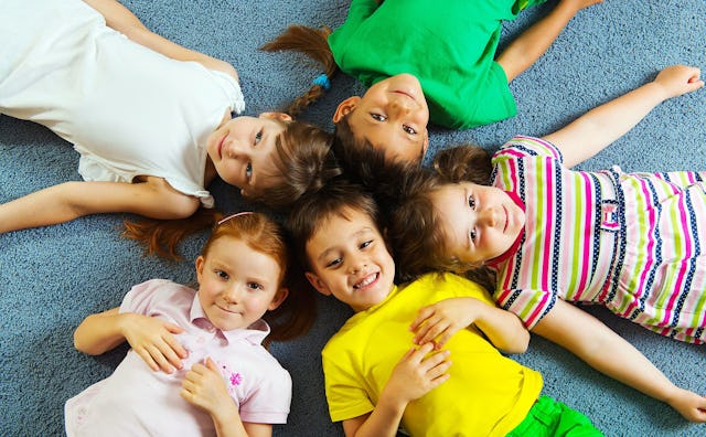Five colorfully dressed children laughing as they lie in a circle on the floor with their heads toge...