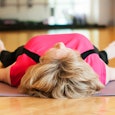 A blonde woman in a pink shirt and black yoga pants lying on a grey yoga mat which is placed on a br...