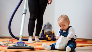 A baby sitting on the floor while his mom is vacuuming around him.