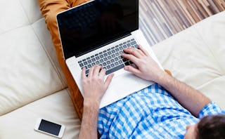 A man in a checked long-sleeved shirt and brown pants typing on his laptop 