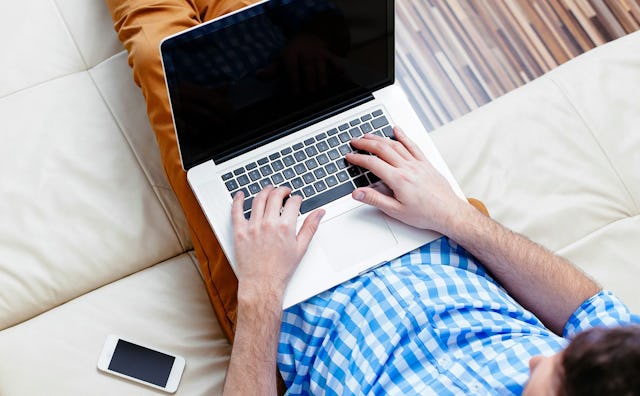 A man in a checked long-sleeved shirt and brown pants typing on his laptop