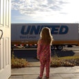 The back of a small girl standing at the entrance of a house waving to a big truck 'United Van Lines...