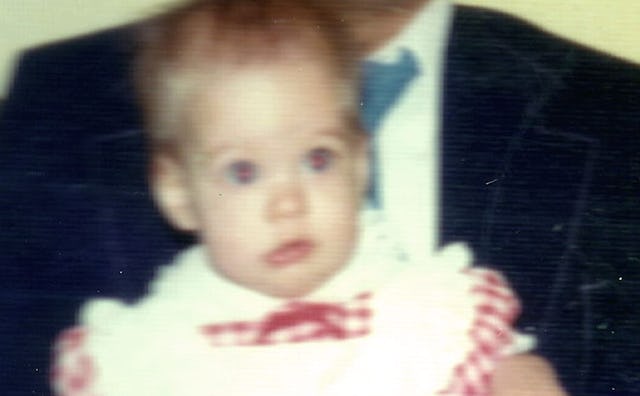 An old blurry picture of a baby in white and red dress being held by her father