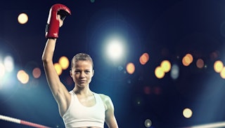 A blonde athletically built woman wearing a white tank top and boxing gloves while holding one hand ...