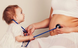 A toddler in a white shirt using a blue stethoscope on her mother's pregnant belly