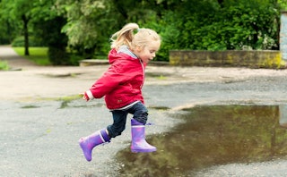 A little girl in a pink jacket and purple boots jumping into a puddle