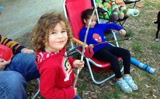 A kid standing and holding a stick next to a girl and a parent sitting on red chair during camping