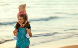 A young brown-haired mother in a blue T-shirt carrying her blonde toddler daughter on her shoulders ...