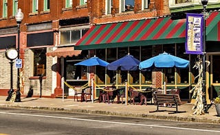 A small-town street view of a brown building and a coffee shop with blue parasols and tables with ch...