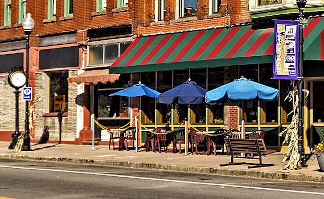 A small-town street view of a brown building and a coffee shop with blue parasols and tables with ch...