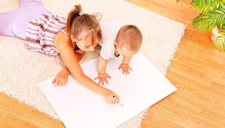 A tween girl drawing with her toddler brother on paper placed on a floor with both of them lying sto...