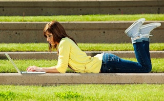 A college girl lying down on grass and working on her laptop 
