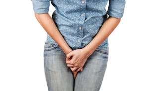 A woman in a blue shirt and gray denim jeans holding her hands together tightly between her legs rep...