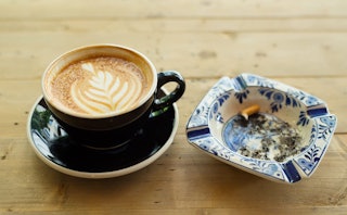 A cup of latte with a coffee art next to an ashtray with blue drawings and one cigarette inside.