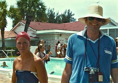A woman and a man posing for a picture in front of a big house with a pool, and a crowd of people.