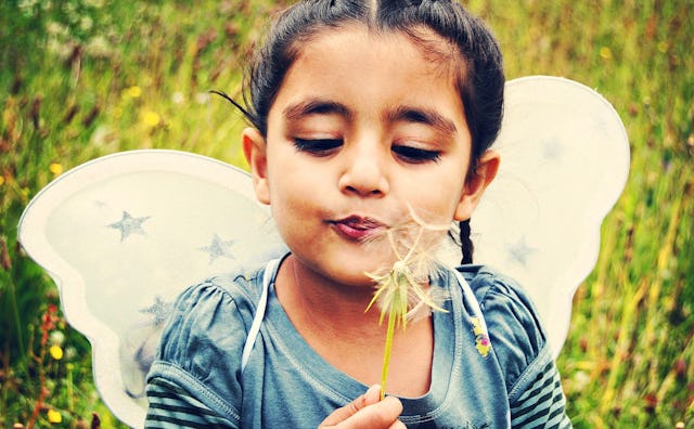 An eight-year-old girl in a blue shirt blowing a dandelion with white fairy wings on her back