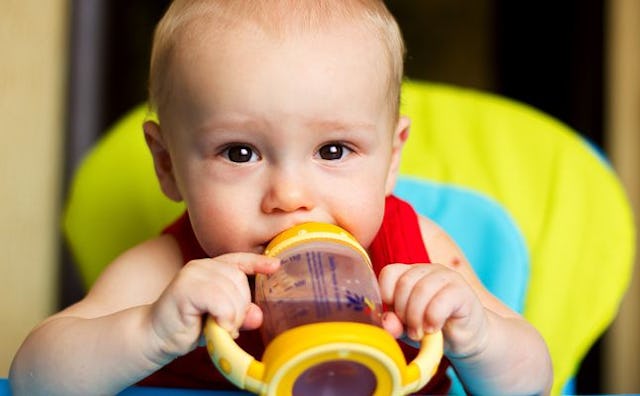 A blonde newborn with watery eyes sitting in a yellow chair and holding his bottle with a brown liqu...