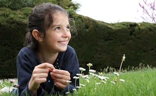 A brown-haired girl in a black shirt lying on the grass and smiling, and next to her are daises.