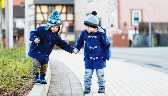 Two toddler boys on a sidewalk, one is being kind by helping the other walk and holding his hand