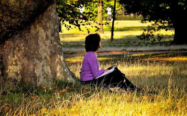 A young woman sitting next to a three while holding a book on her lap