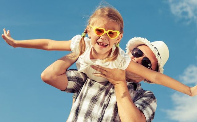 A dad in a plaid shirt and straw hat holding his daughter up while she spreads out her arms as if fl...