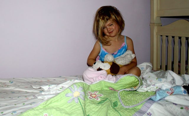 A girl with a bob haircut in a blue tank top lying in bed holding her plush toy with a white-green f...