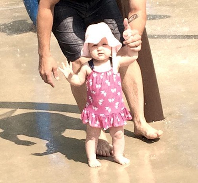 A baby girl with a hat on walking on the sand while a person is holding one of her arms.