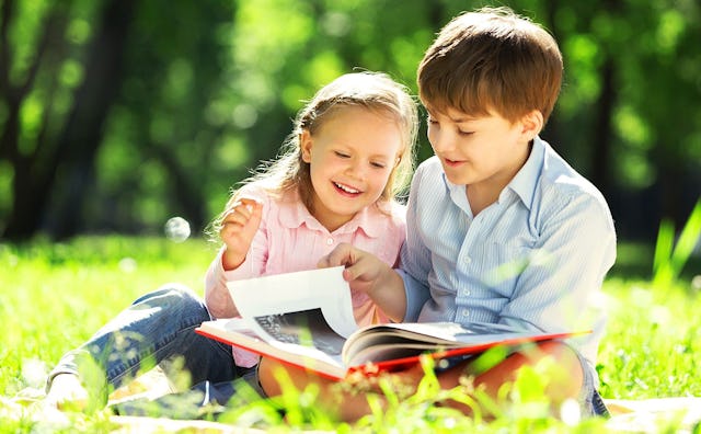 A young girl and a boy sit beside each other in a meadow while reading a book
