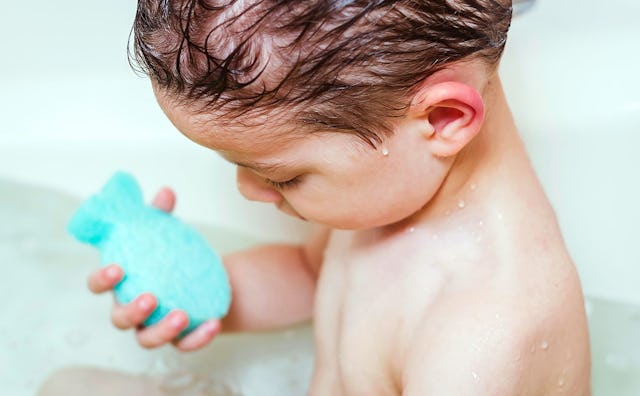 A brown-haired toddler boy taking a bath and holding a blue fish-shaped sponge getting his hair wash...