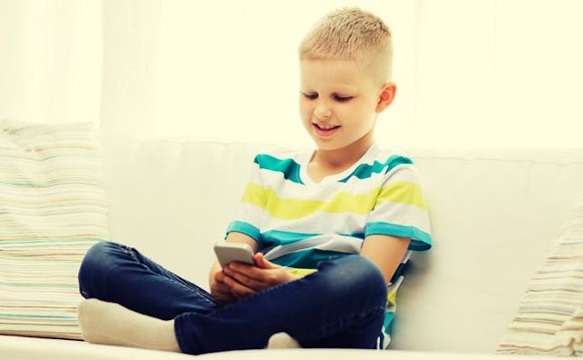 A 10-year-old boy is sitting on a couch, looking at a cell phone and smiling