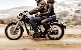 A man in a brown leather jacket and a woman in a black leather jacket riding a motorcycle/blurred du...