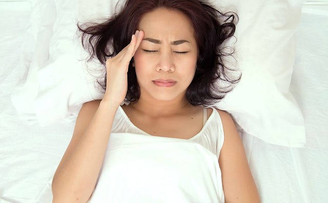 Young hungover mom lying in bed with white sheets, holding her arm on her head with her eyes closed