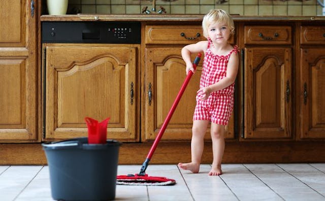 A little blonde girl holding a mop, ready to help her mum clean, while wearing a red and white rompe...