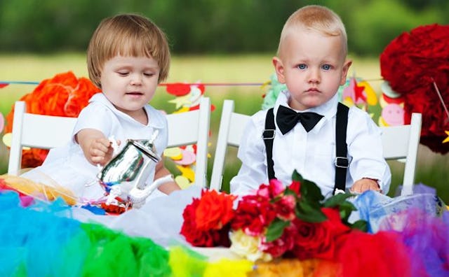 Two toddlers sitting at a white table with floral decorations while the girl is sipping tea