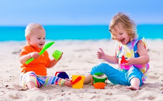 A baby and a 2-year-old child playing on the beach with colorful toys and smiling