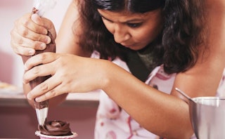 A dark-haired woman wearing an apron while squeezing out chocolate topping on a muffin in a kitchen