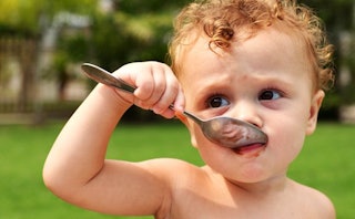 A toddler eating with a large spoon with a blurred park in the background