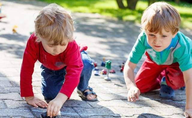 Two kids playing on the ground, and one of them is drawing something with chalk.