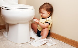 A toddler sitting next to the closed toilet seat in a public restroom, unwrapping the toilet paper