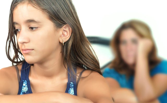 A sad tween girl looking down, with her mother looking at her in the background.