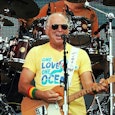Jimmy Buffett playing the guitar in a yellow shirt saying "One Love One Ocean".