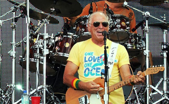 Jimmy Buffett playing the guitar in a yellow shirt saying "One Love One Ocean".