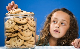 A small girl opens up a glass jar full of cookies and takes one out